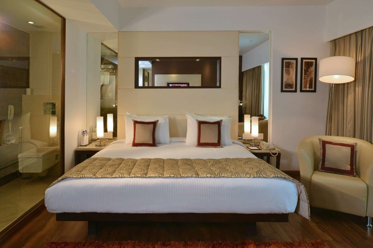 Habitación de hotel con una cama grande, un espejo sobre la cama, una silla, un escritorio con lámpara y un tapete rojo.