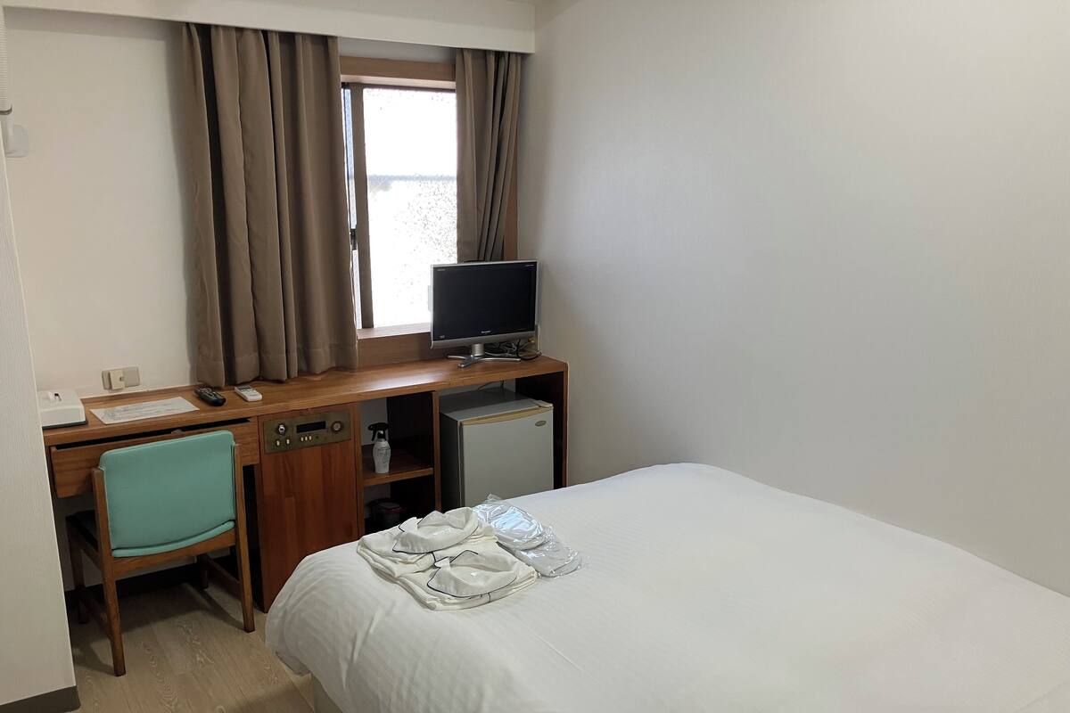 Une chambre d’hôtel comprenant un lit, un bureau avec un ordinateur, une chaise, une fenêtre avec des rideaux et un petit réfrigérateur.