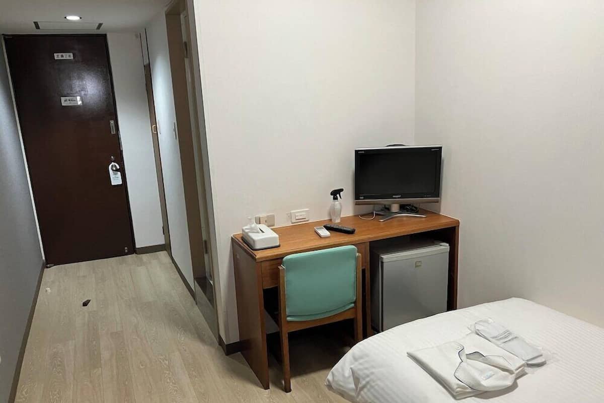 A hotel room with a bed, a desk with a computer, a chair, a small fridge, and a door.