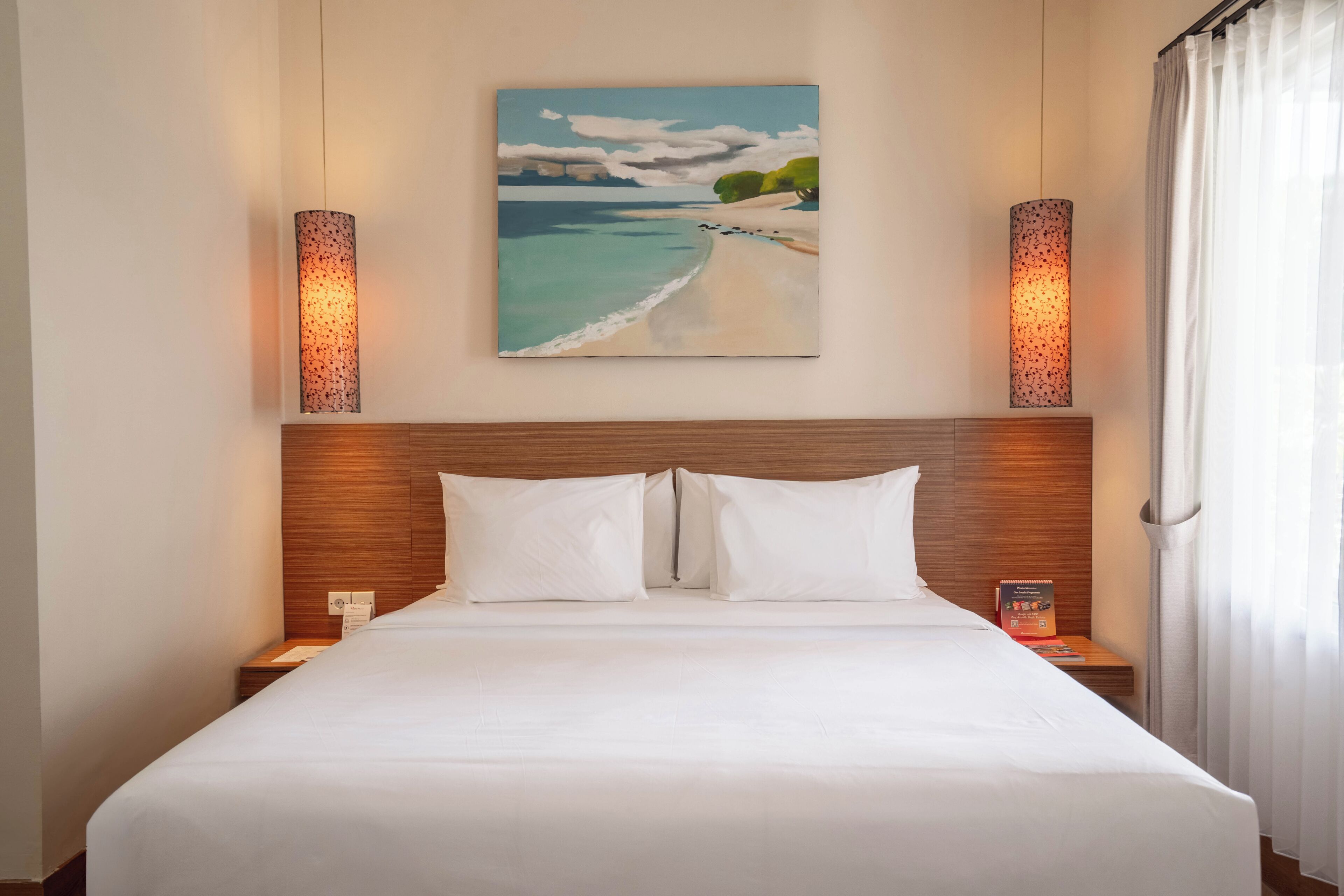 A hotel room with a large bed, two bedside tables, a nightstand, a painting of a beach scene, and two wall-mounted lamps.