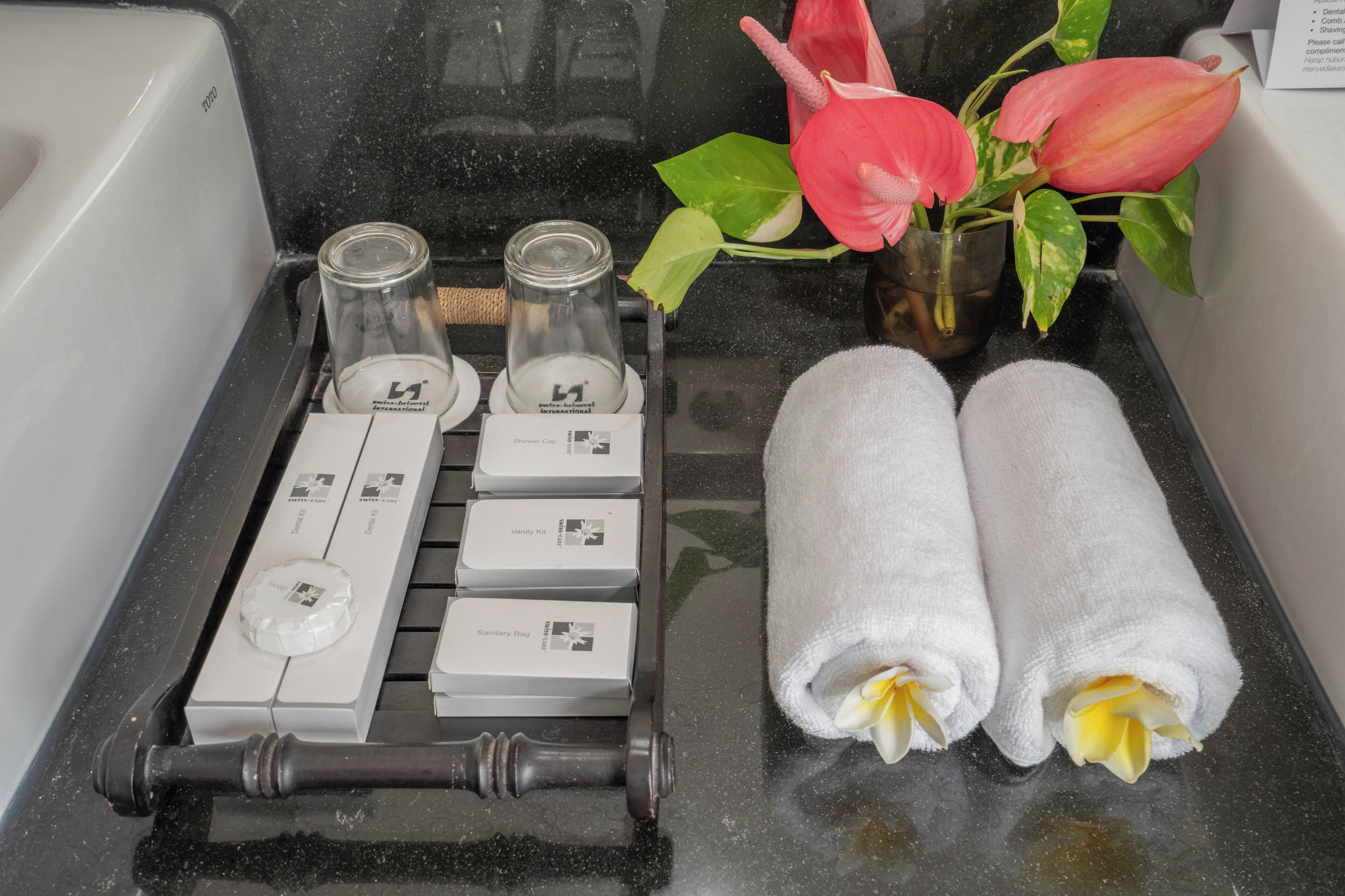 Ett badrum i ett hotellrum med en hylla ovanför handfatet, där toalettartiklar står, en handduksställning och en vas med blommor.