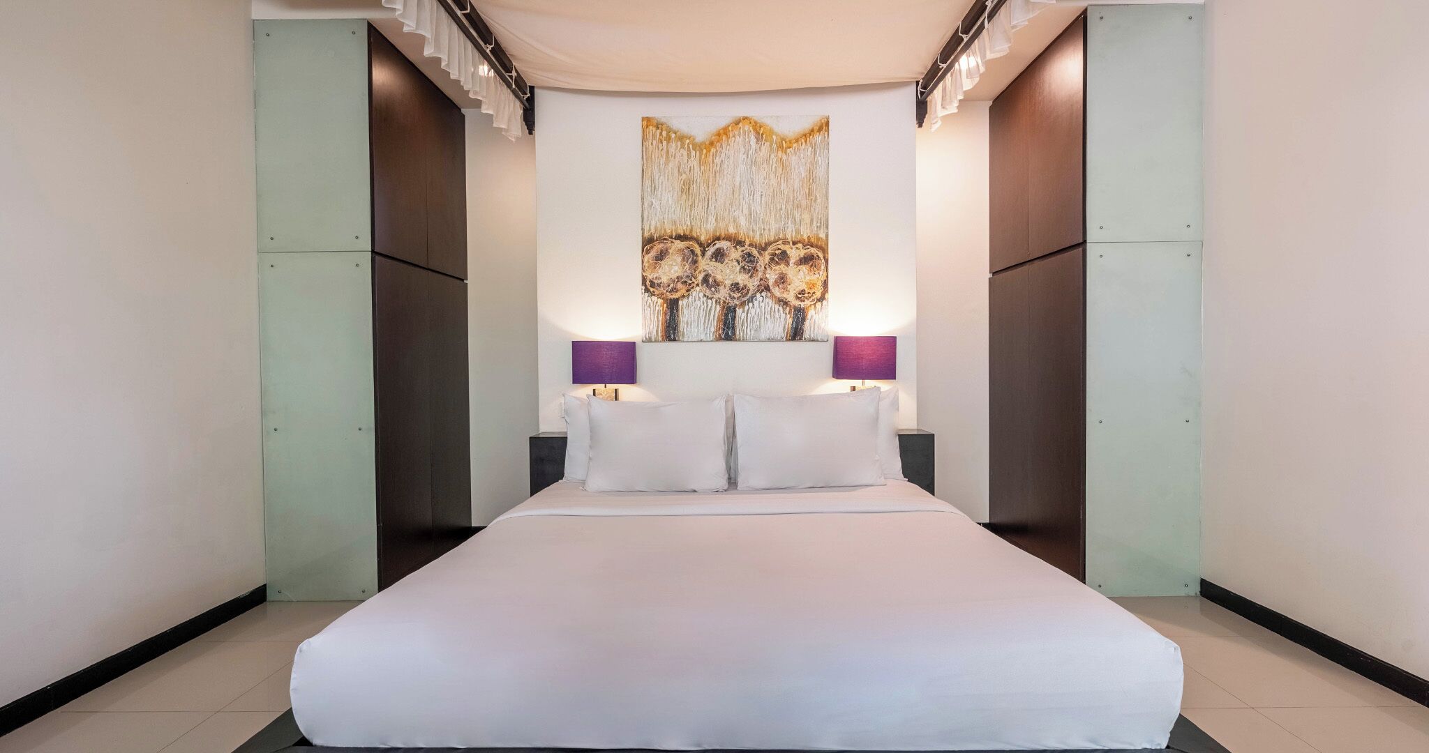 A large bed with white linens, two bedside lamps, and a decorative wall hanging above.