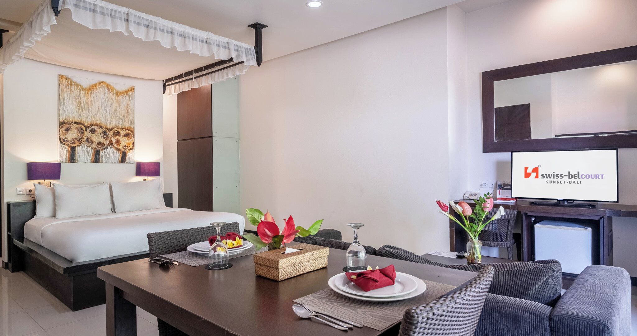 Habitación de hotel con cama, una mesa de comedor con platos y cubiertos, un televisor y un cuadro grande en la pared.