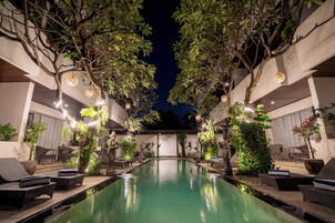 Uma área de piscina com espreguiçadeiras e plantas em vasos, cercada por árvores e iluminada por luzes penduradas.