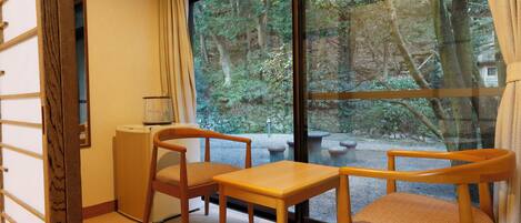 A room with a view of a forest through a large window, a small table, two chairs, and a lamp.