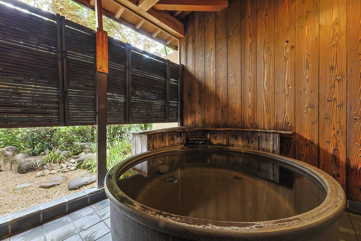 A traditional wooden bath with a stone exterior, surrounded by a wooden fence and a view of a garden.