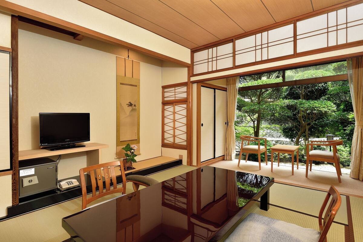 A traditional Japanese-style room with tatami flooring, a low table, a television, and a view of a garden through large windows.