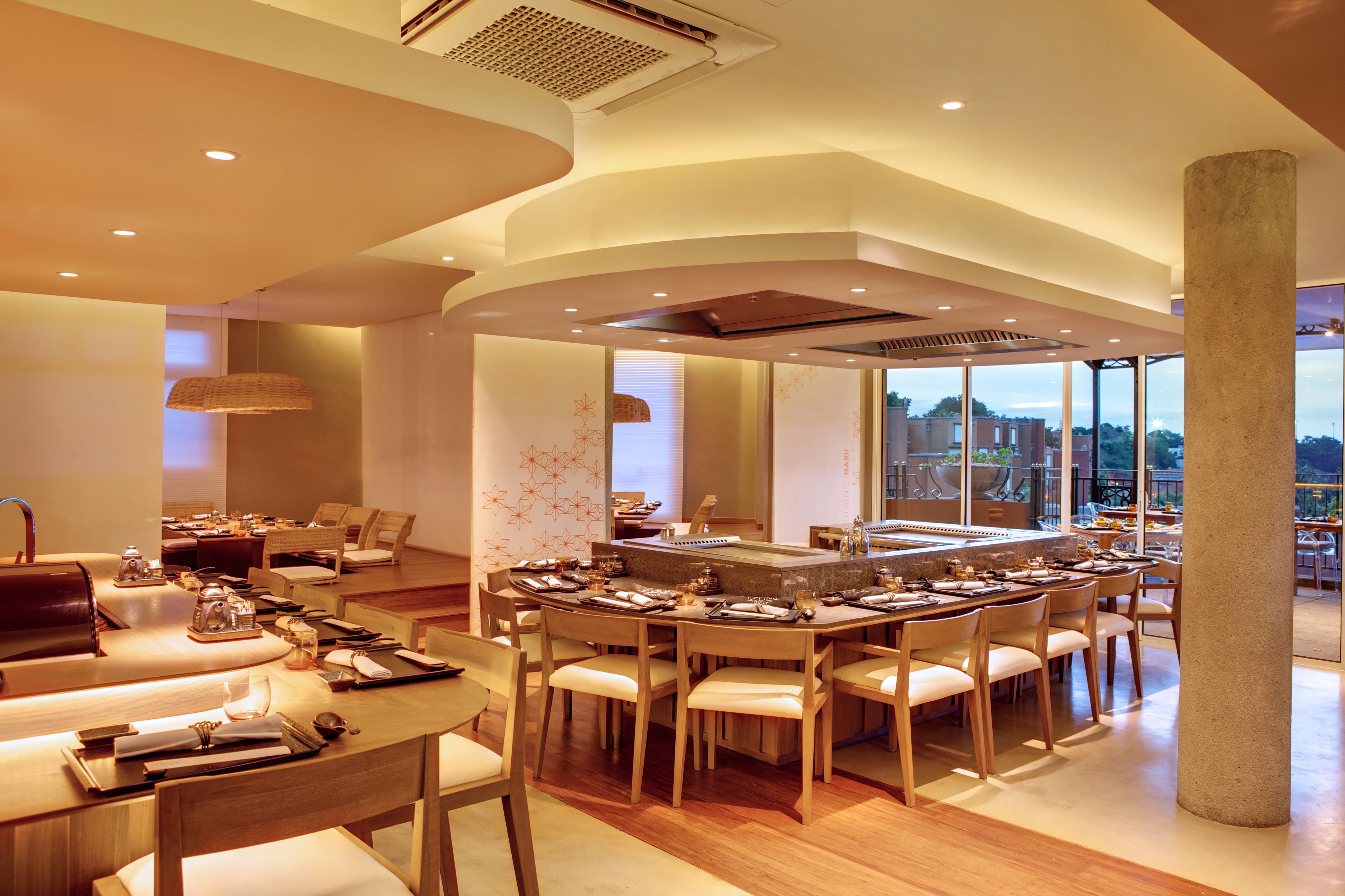 A modern dining area with multiple tables and chairs, a central bar, and large windows offering a view of the cityscape.