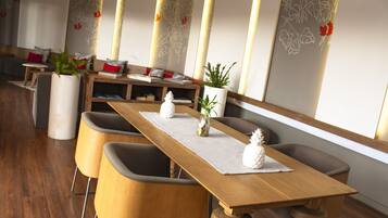 A modern dining area with wooden tables and chairs, potted plants, and books on shelves.