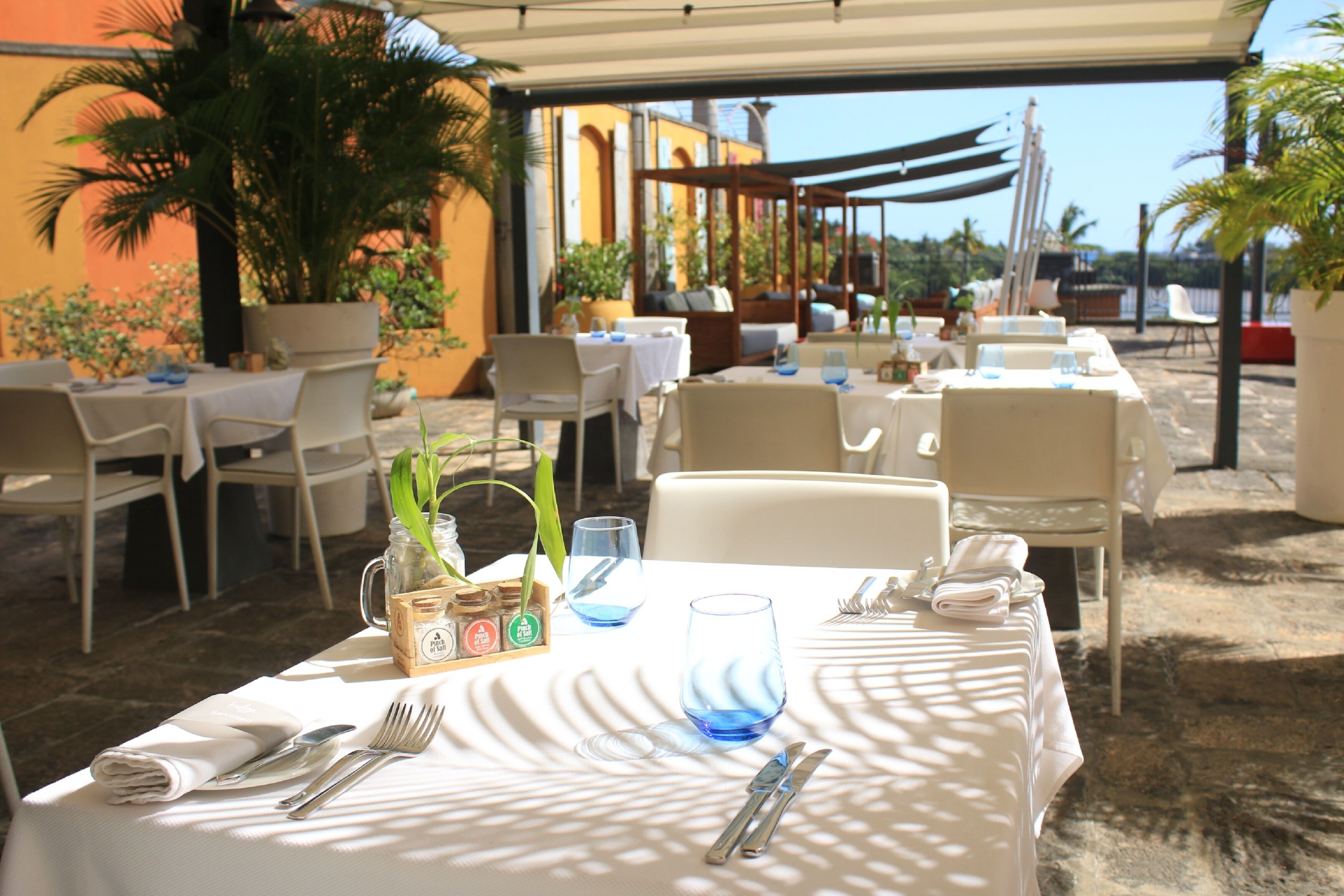 Un área de comedor al aire libre con mesas preparadas para dos, con manteles blancos, servilletas dobladas con cuidado y cubiertos.