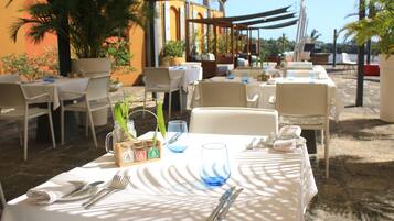 Un área de comedor al aire libre con mesas preparadas para dos, con manteles blancos, servilletas dobladas con cuidado y cubiertos.