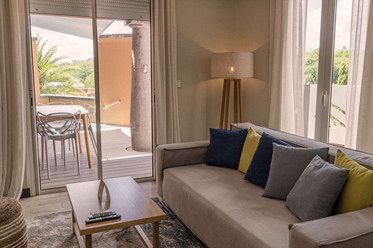 A living room with a sofa, a coffee table, and a lamp. There is a sliding glass door leading to an outdoor area with a table and chairs.