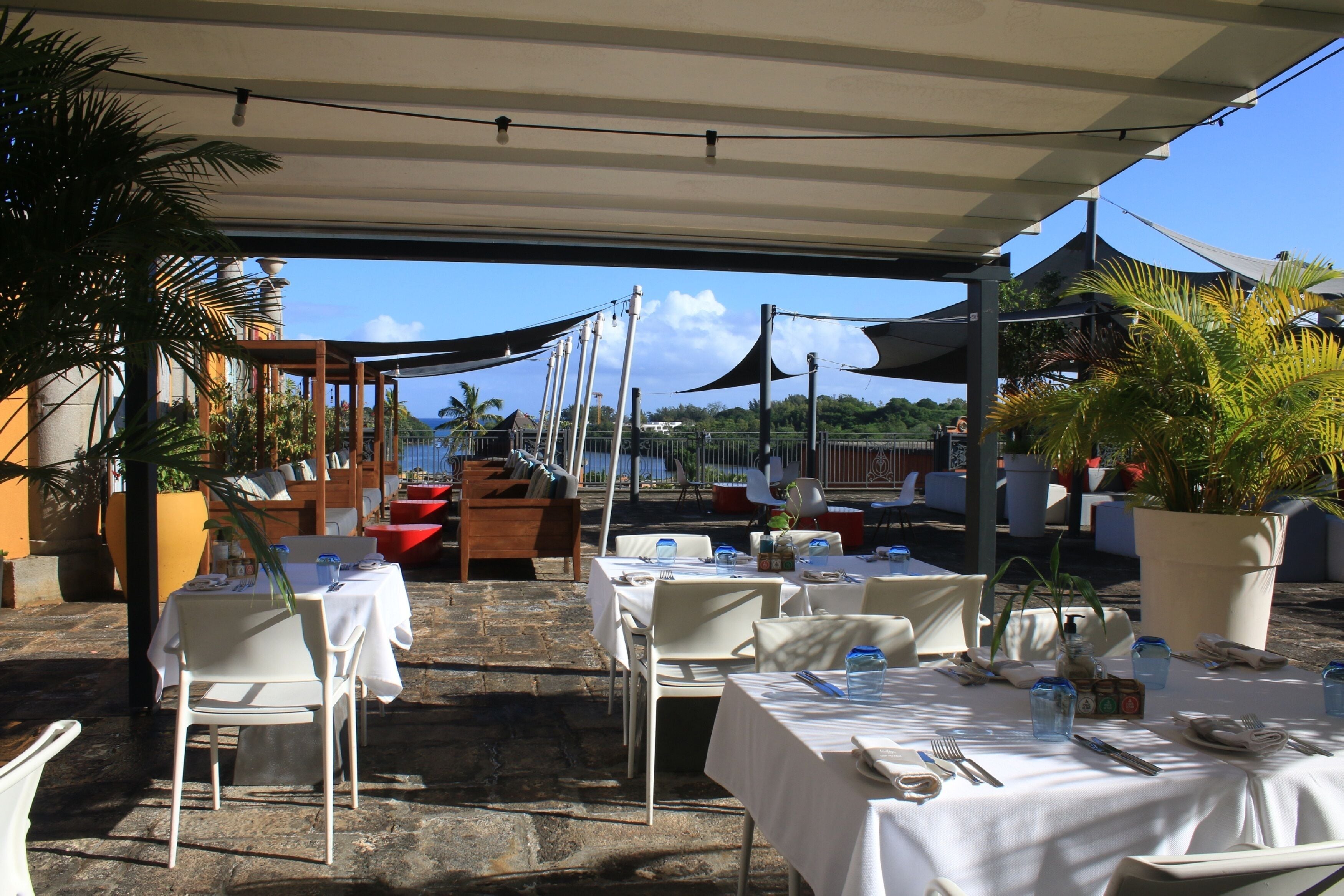 An outdoor dining area with tables and chairs, covered by a canopy, and surrounded by potted plants.