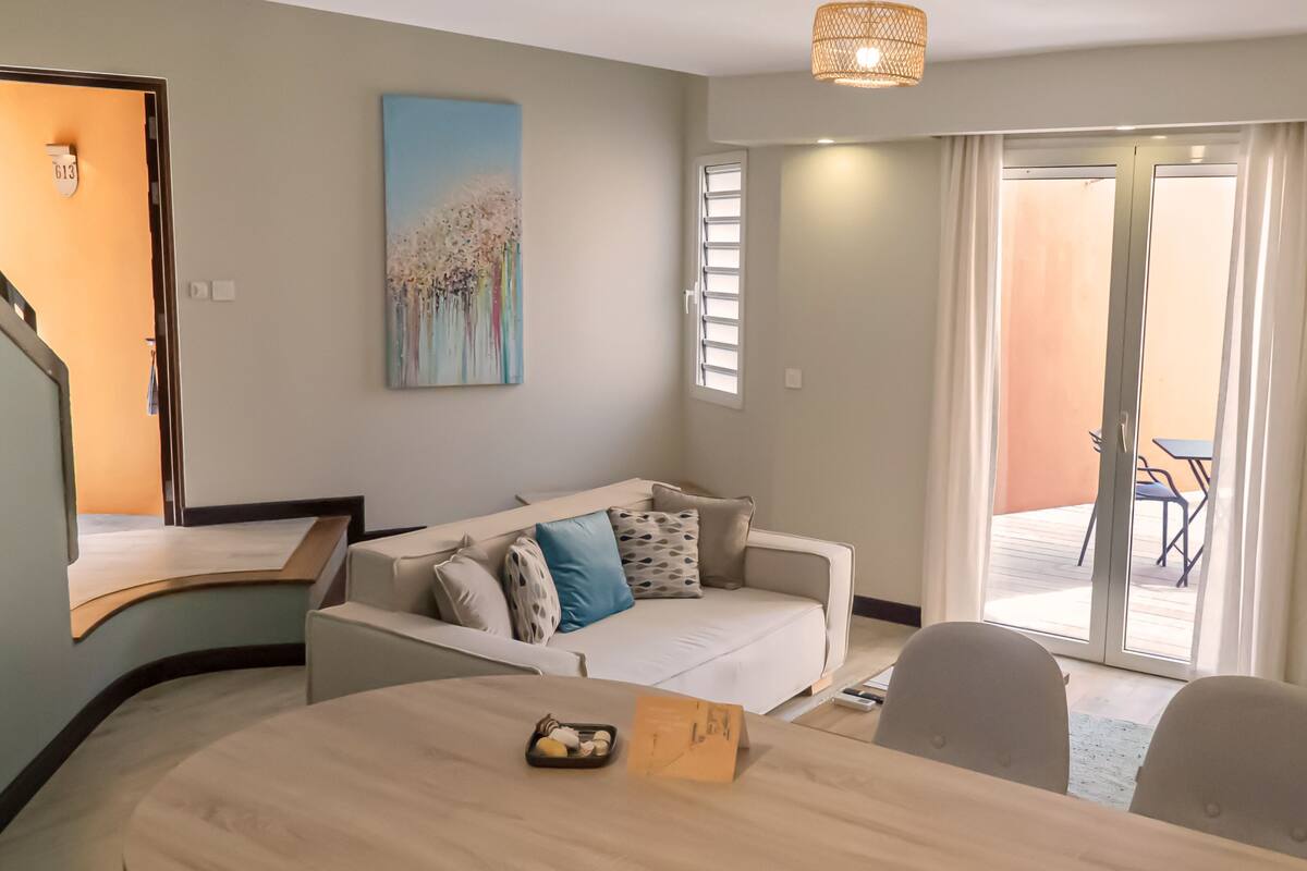 A modern living room with a sofa, a dining table with a tray of snacks, a painting on the wall, and a view of the outdoor patio area.