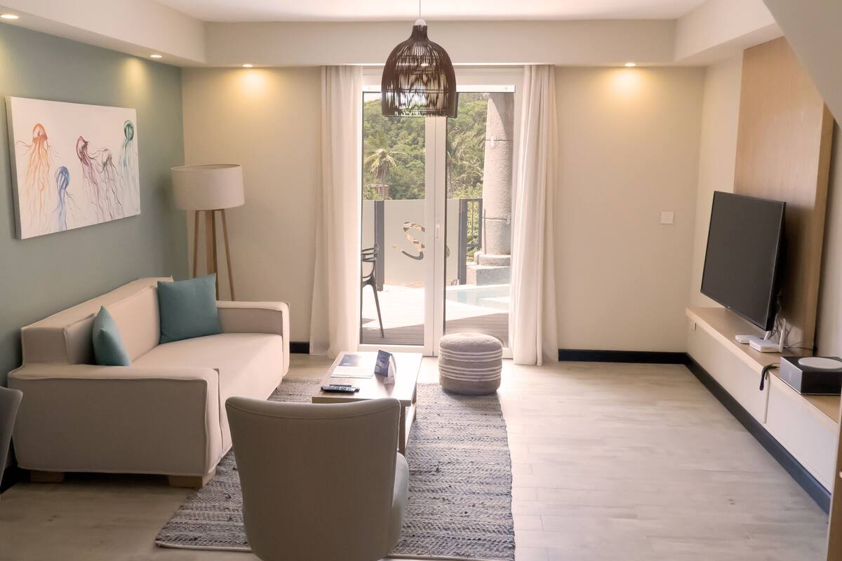 A modern living room with a sofa, coffee table, and TV. There is a painting on the wall, a lamp, and a view of the outdoors through a glass door.