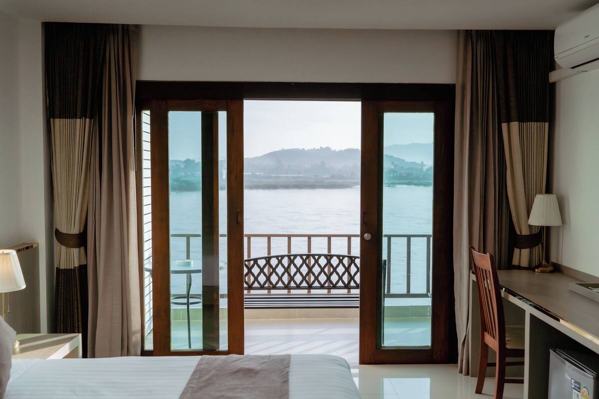 Habitación de hotel con cama, escritorio, silla, televisor y balcón con vistas a un cuerpo de agua y colinas.