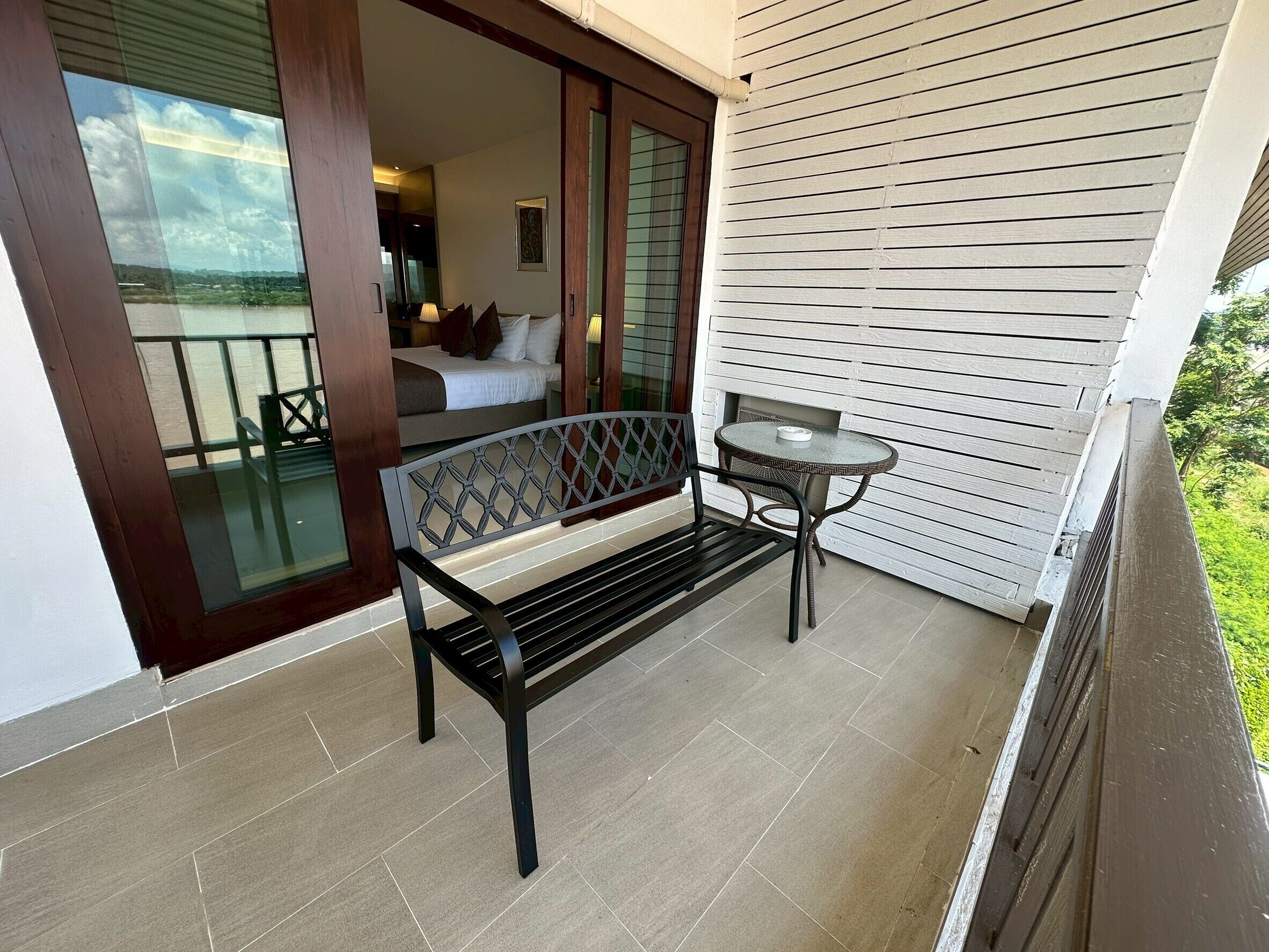 A balcony with a bench and a small table, a sliding glass door leading to a bedroom, and a view of the outdoors.