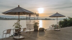 Una terraza con mesas y sillas, dos sombrillas, un portón metálico y vistas al agua y a las montañas.