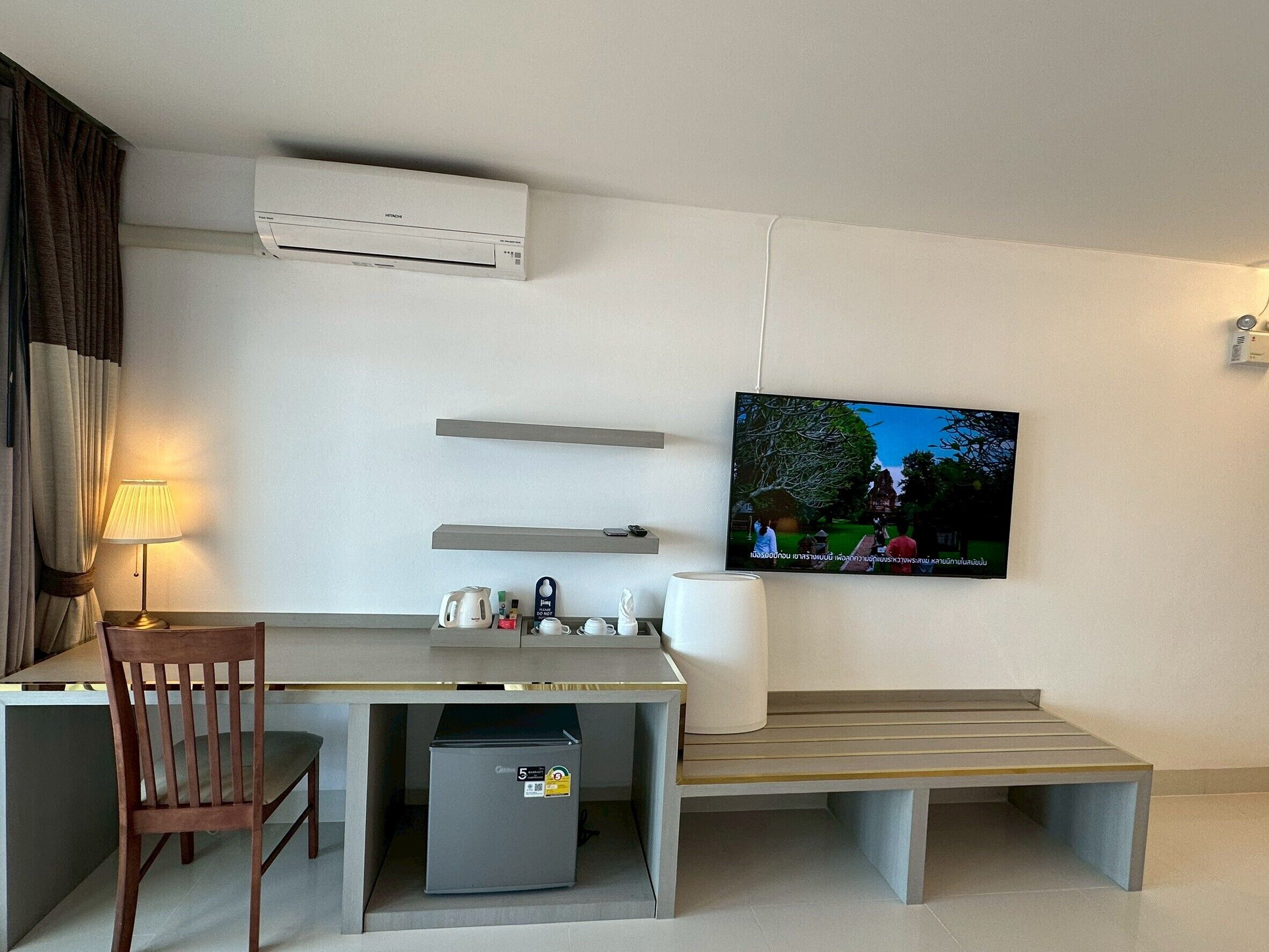 Une chambre d’hôtel équipée d’un bureau, d’une chaise, d’une banquette, d’un téléviseur, d’un climatiseur et d’un petit réfrigérateur.