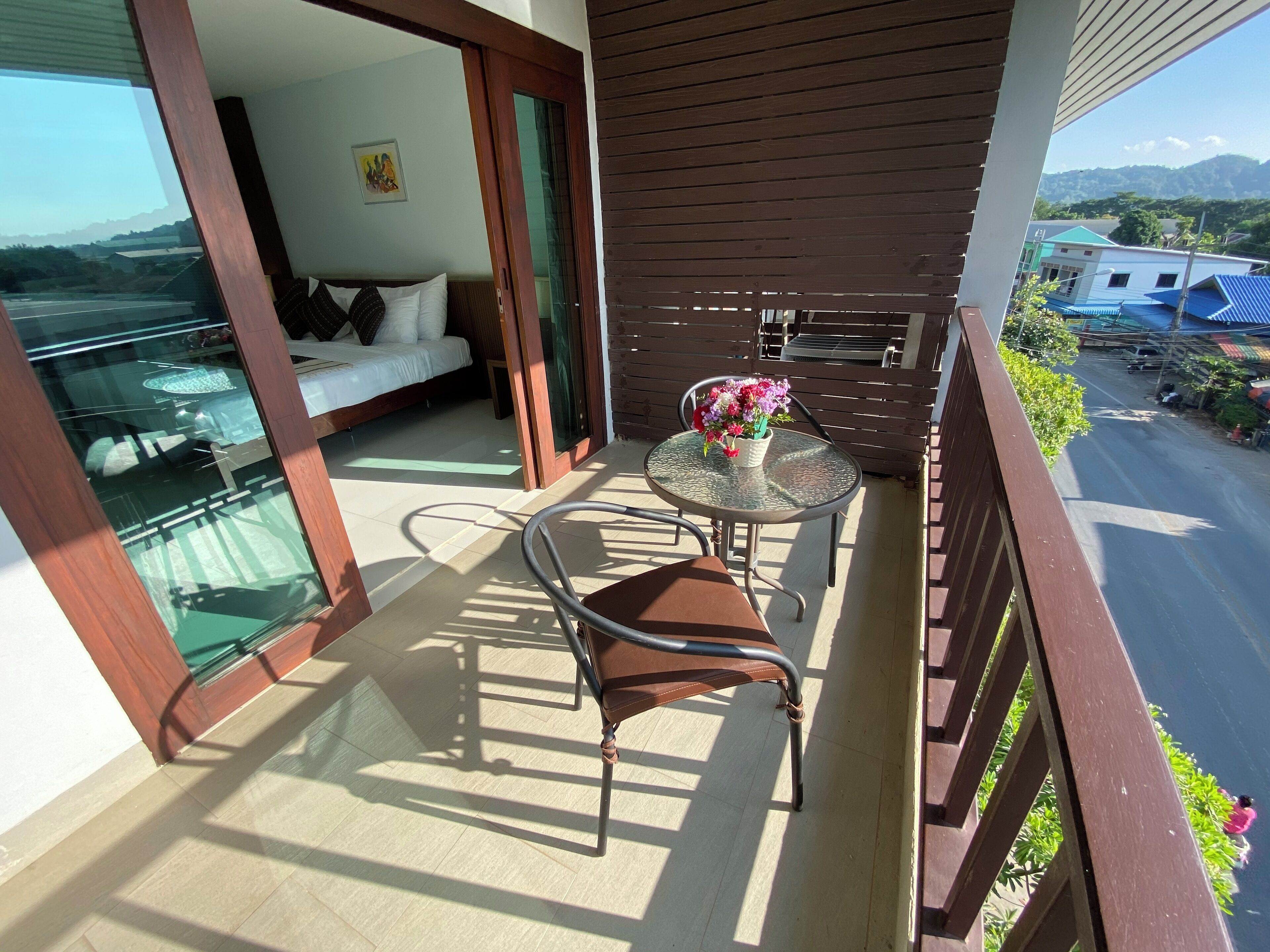 A balcony with a bed, a table with flowers, and a view of the road and buildings.