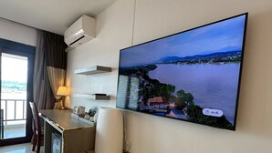 Habitación de hotel con un televisor de pantalla plana grande montado en la pared, mostrando una vista panorámica de un lago y montañas. Cuenta con un escritorio, una lámpara, una silla y una mesita con una taza y un control remoto.
