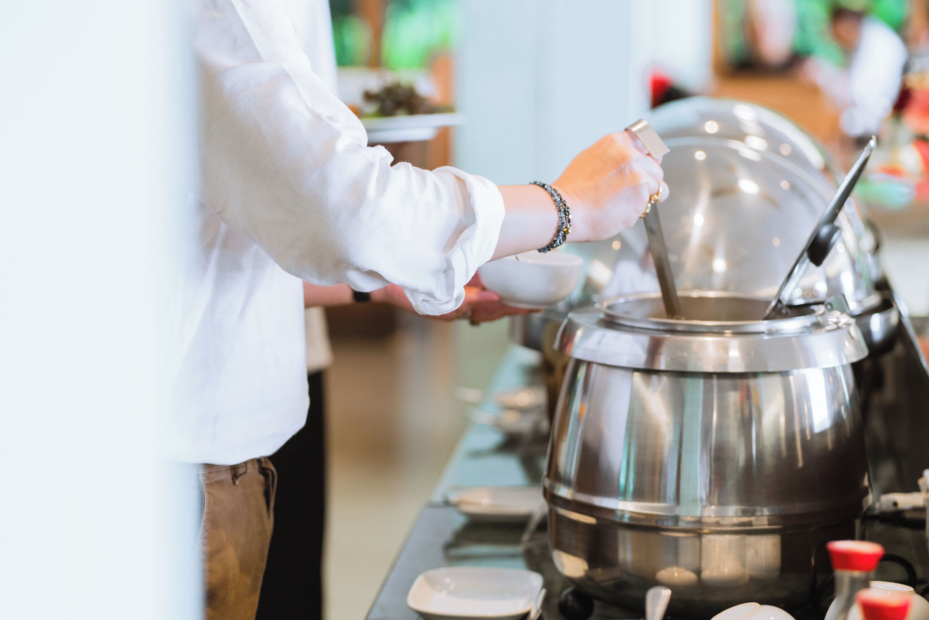 A buffet setup with a large stainless steel pot, a person serving food, and various dishes on the counter.