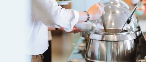 A buffet setup with a large stainless steel pot, a person serving food, and various dishes on the counter.