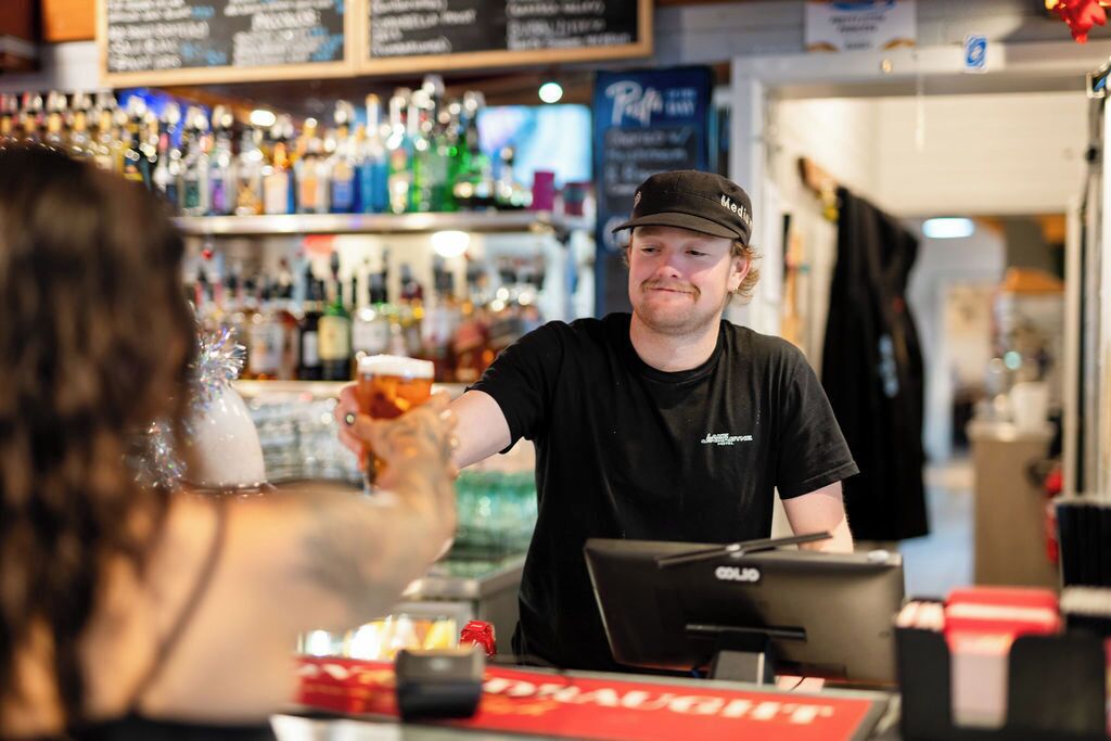 A bar setting with a bartender, a cash register, and a variety of bottles in the background.