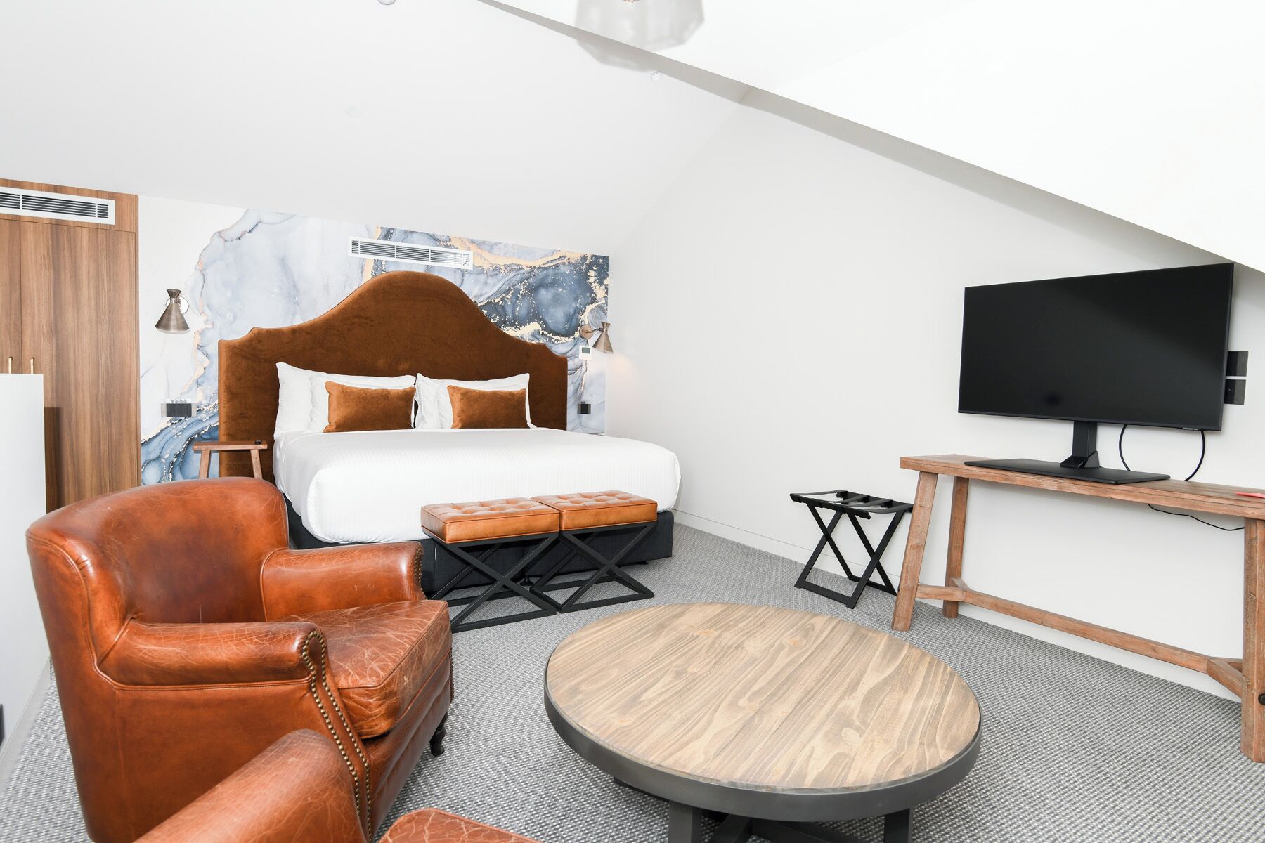 A modern hotel room with a large bed, leather armchair, round wooden table, and a flat-screen TV.