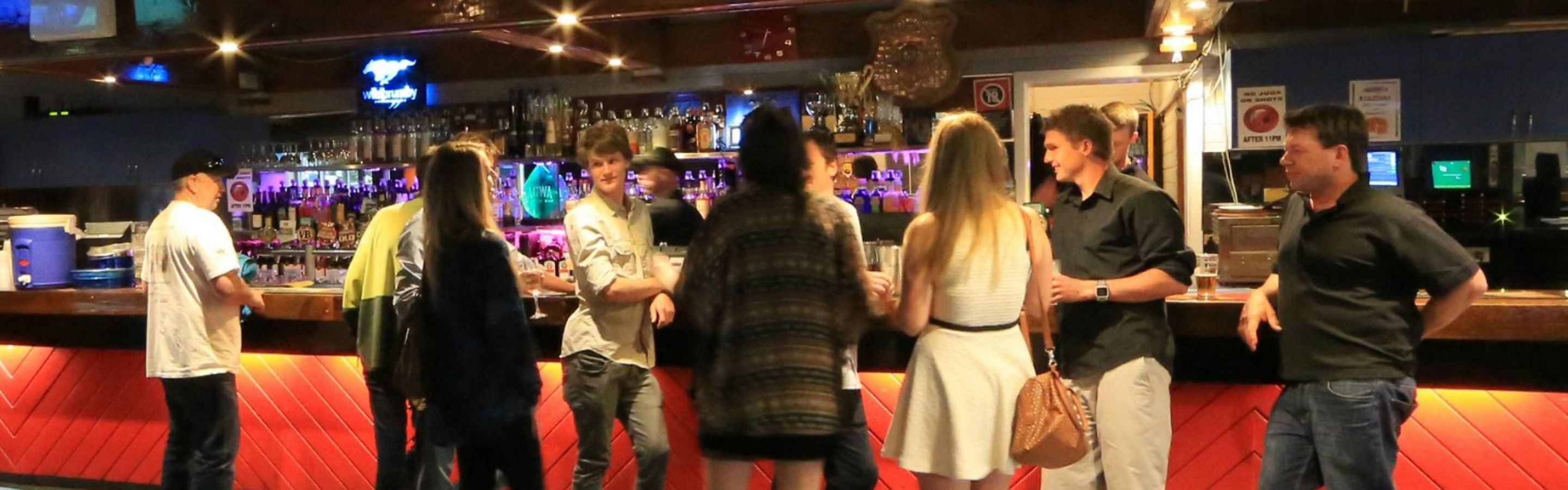 Una zona de bar con una zona de estar acolchada de color rojo, una barra con diferentes botellas y un letrero de neón al fondo.
