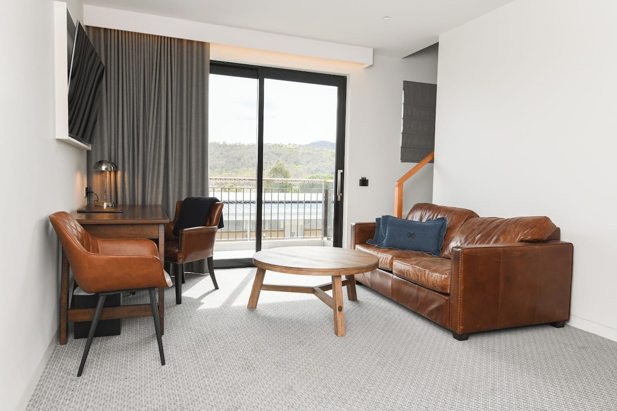 Un salon moderne avec un canapé en cuir marron, une table basse en bois et un fauteuil en cuir marron. On y trouve un bureau avec une lampe, et une grande porte coulissante offre une vue sur l’extérieur.