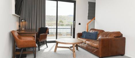 A modern living room with a brown leather sofa, a wooden coffee table, and a brown leather chair. There is a desk with a lamp and a view of the outdoors through a large sliding door.