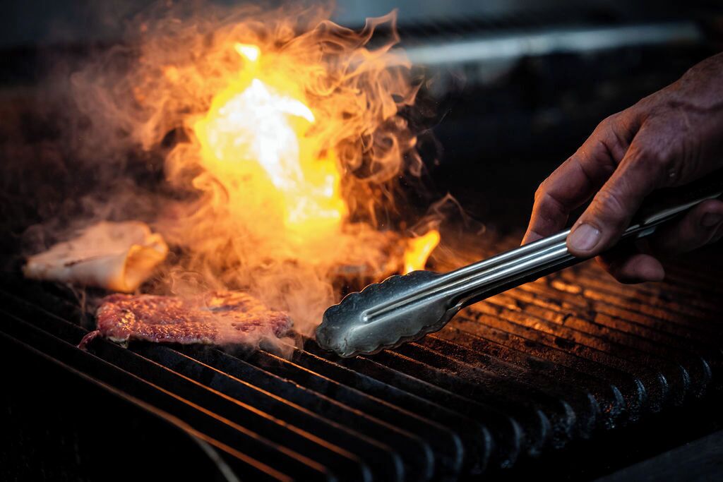 A hand using tongs to grill meat on a grill with visible flames.