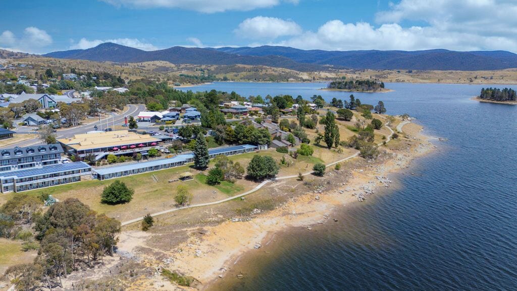 Vue aérienne d’une propriété au bord d’un lac, avec des bâtiments, une plage et des collines environnantes.