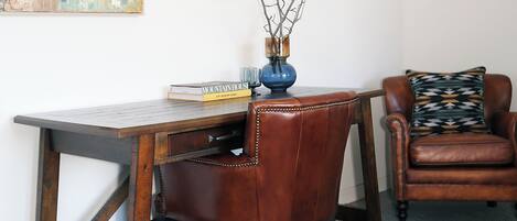 A room with a wooden desk, a leather chair, a vase with branches, a book titled "MOUNTAIN HOUSE," and a colorful abstract painting on the wall.