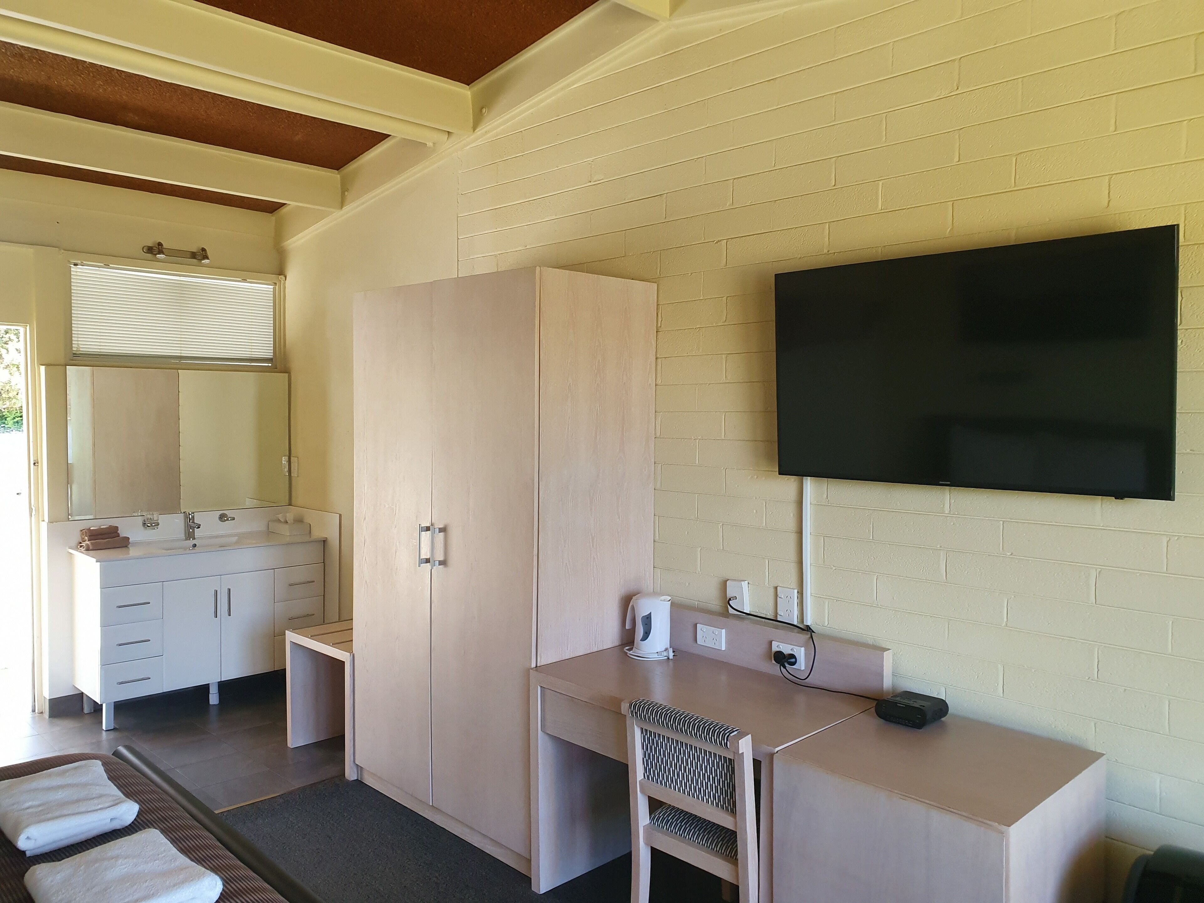A hotel room with a wooden wardrobe, a desk with a chair, a flat-screen TV mounted on the wall, and a large mirror above the vanity.