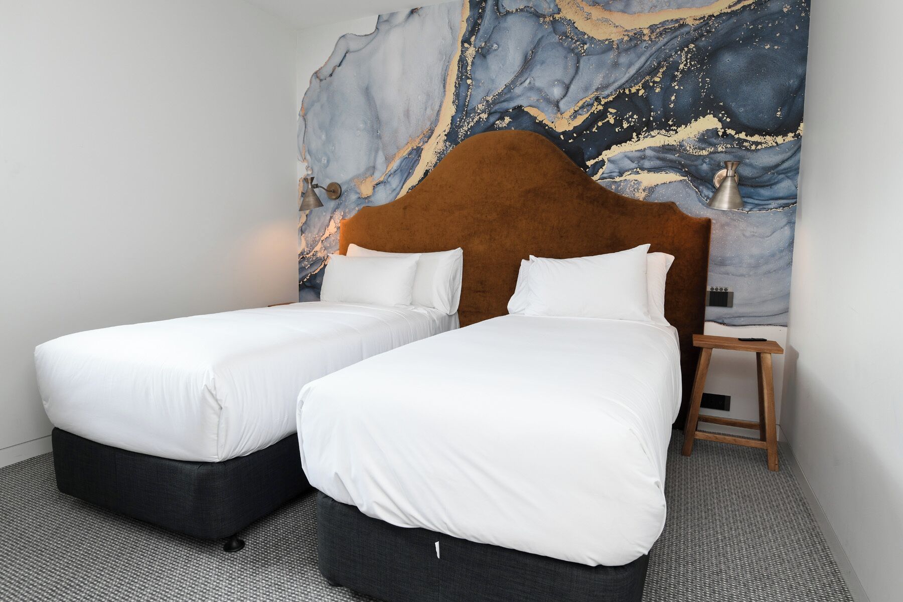 A hotel room with two single beds, a marble wall mural, a wooden nightstand, and a small table.