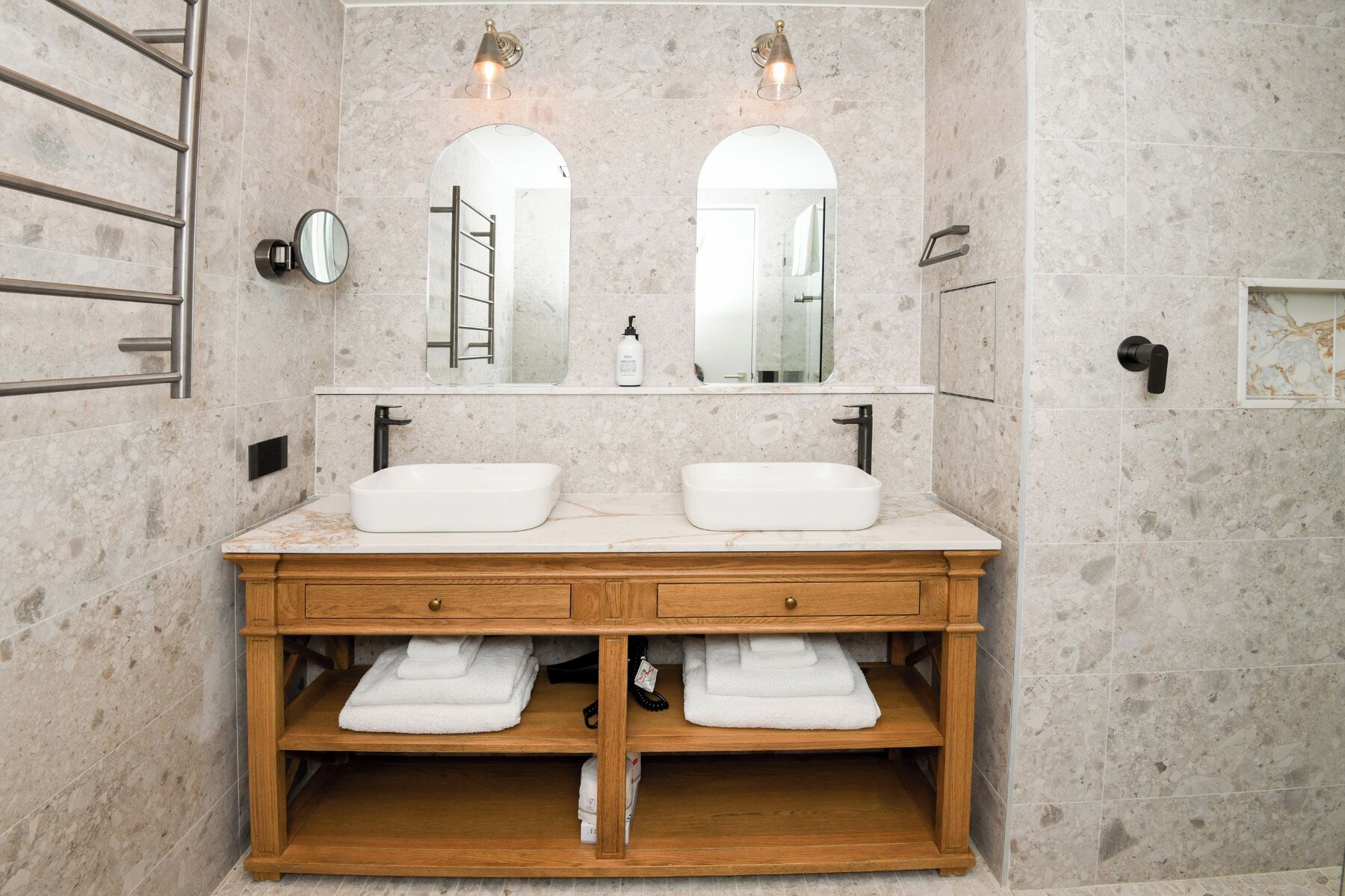 Ein Badezimmer mit zwei Waschbecken, einem Holzbadezimmer-Möbelkorpus, Marmorwänden und zwei Wandleuchten.