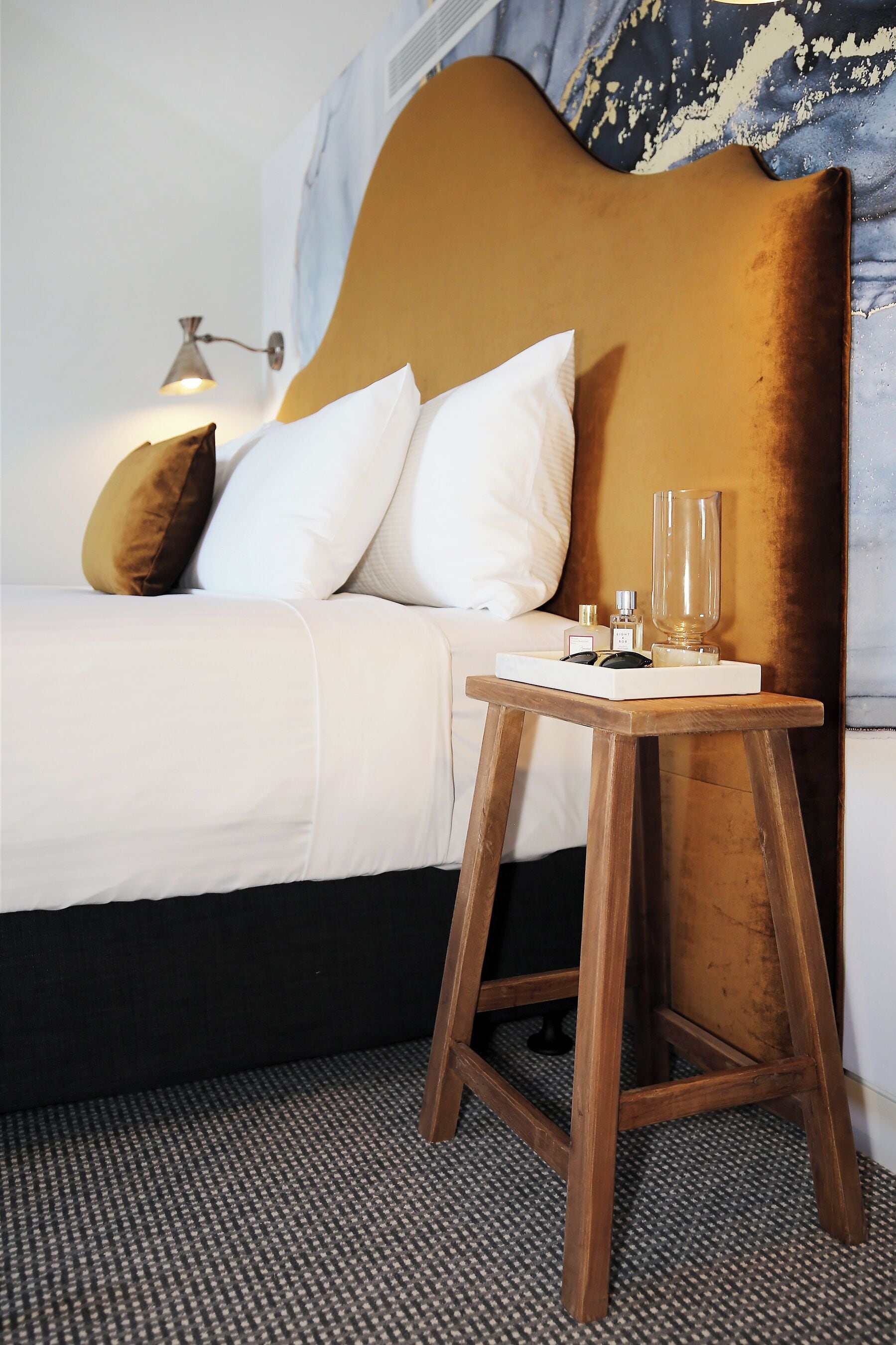 Un lit bien fait, avec du linge de lit blanc, une tête de lit en bois et une table de chevet sur laquelle se trouvent un verre et deux bouteilles.