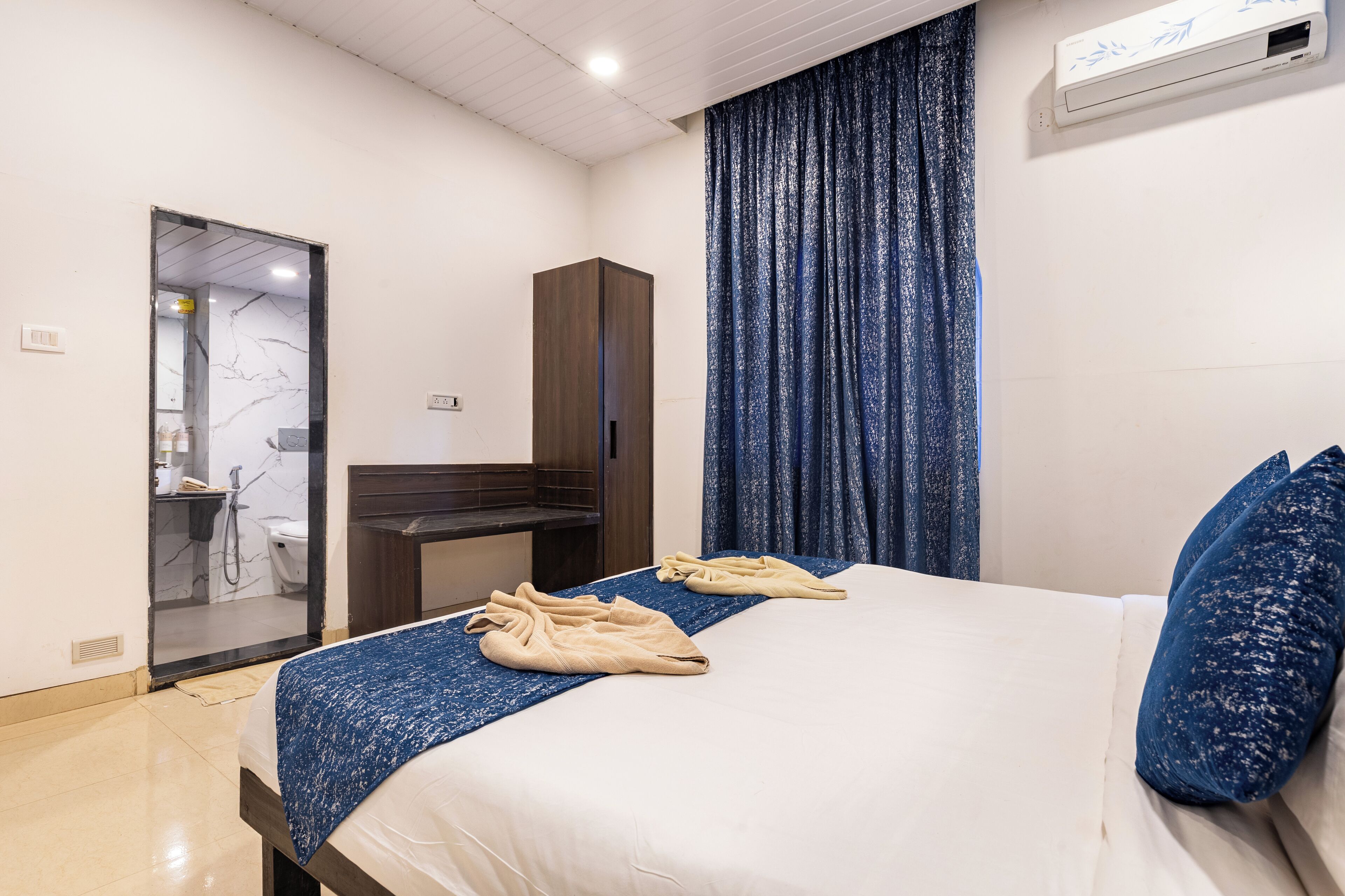 A hotel room with a bed, a desk, a wardrobe, and a bathroom visible through a glass door.