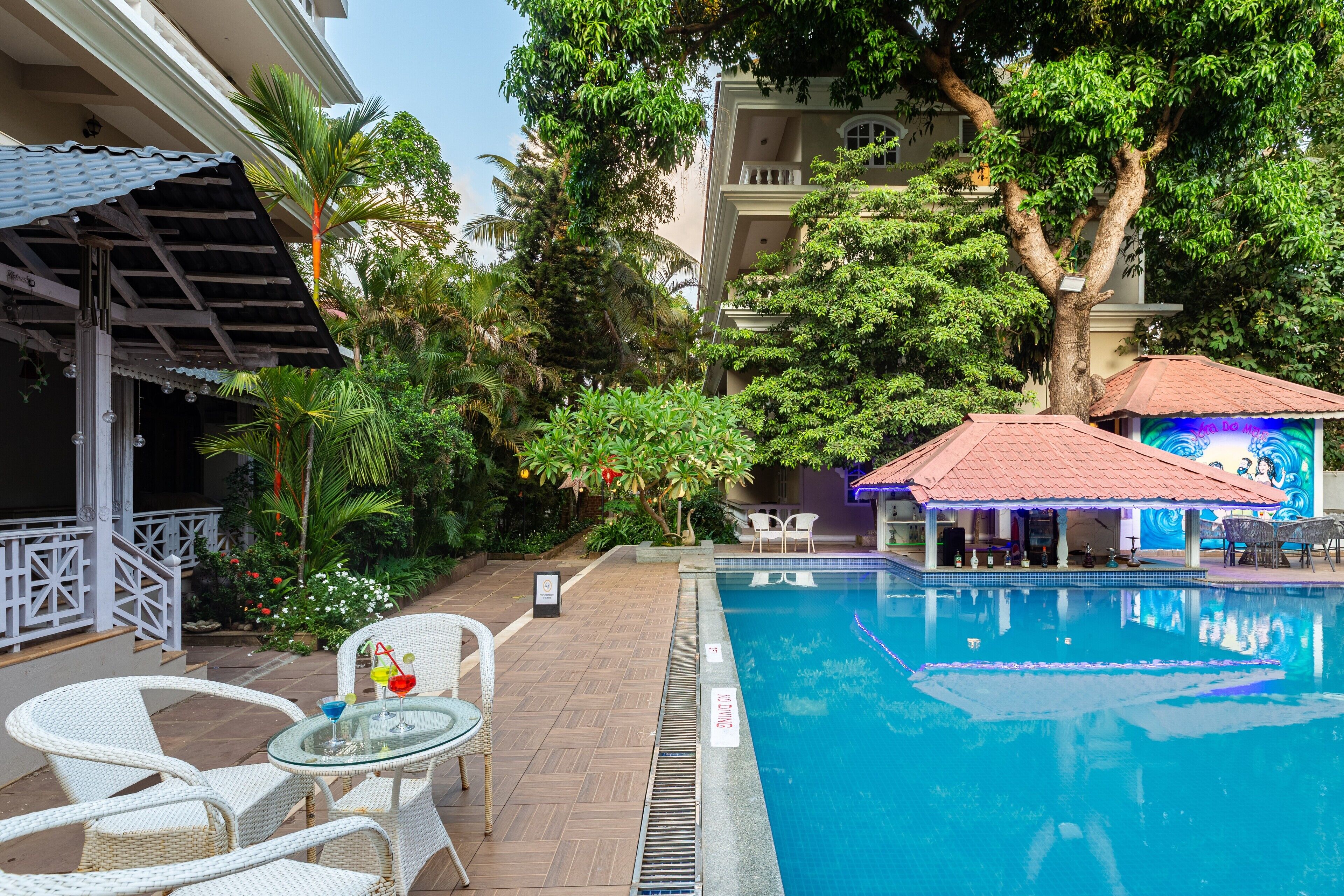 A pool area with a clear pool, white wicker chairs, and a covered seating area with a mural.