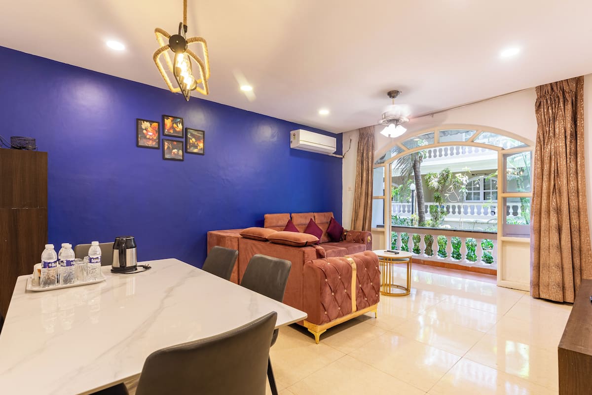 A room with a blue wall, a bed, a dining table with chairs, and a window with a view of greenery.