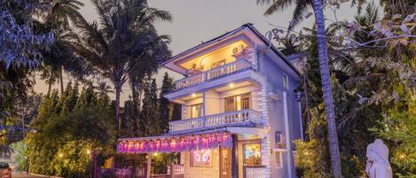 A two-story building with a balcony, surrounded by tropical vegetation.