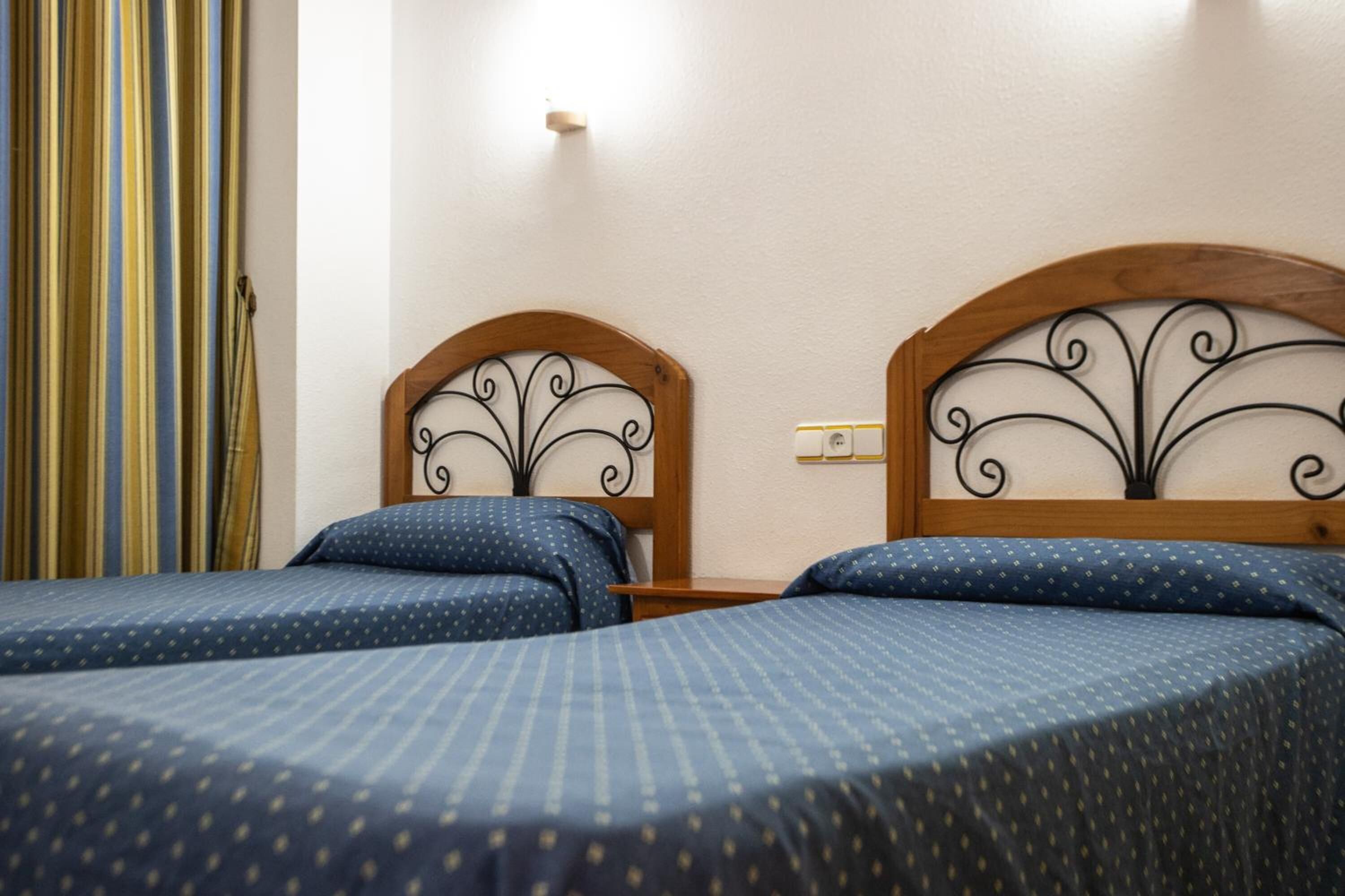 Habitación de hotel con dos camas, cada una con cabecero de madera y cubrecama azul con estampado.