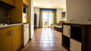 Een compacte keuken met gele kastjes, een ingebouwde oven en een koelkast. Er is een zithoek met een tafel en stoelen, en een groot raam met gordijnen.