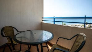 Een balkon met een ronde glazen tafel en twee rieten stoelen, met uitzicht op een strand met een blauwe reling.