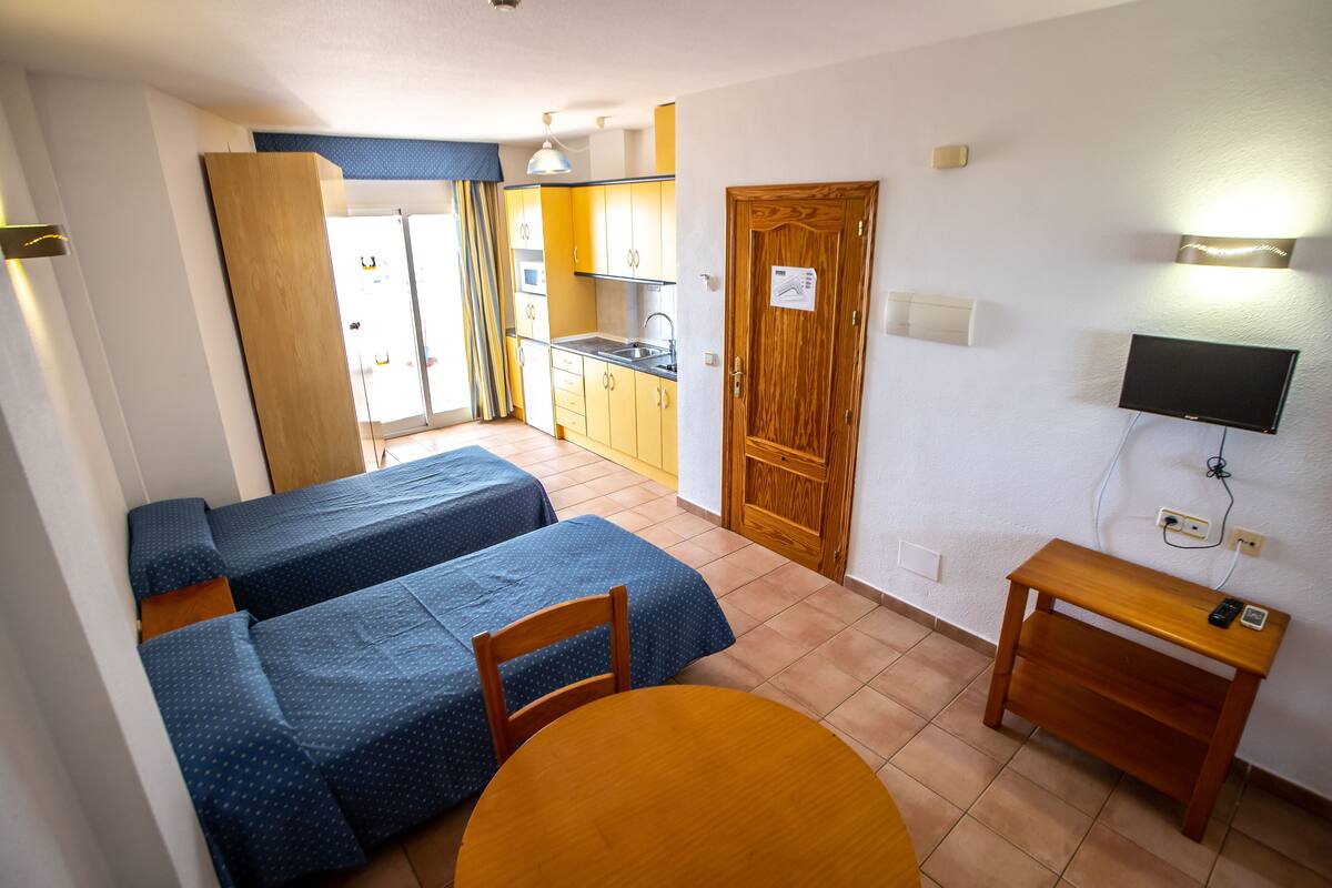 Habitación compacta con dos camas, una mesa de madera, una pequeña cocina y un televisor en la pared.