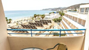 Een balkon met een glazen tafel en rieten stoelen, met uitzicht op een strand en hotelgebouwen.