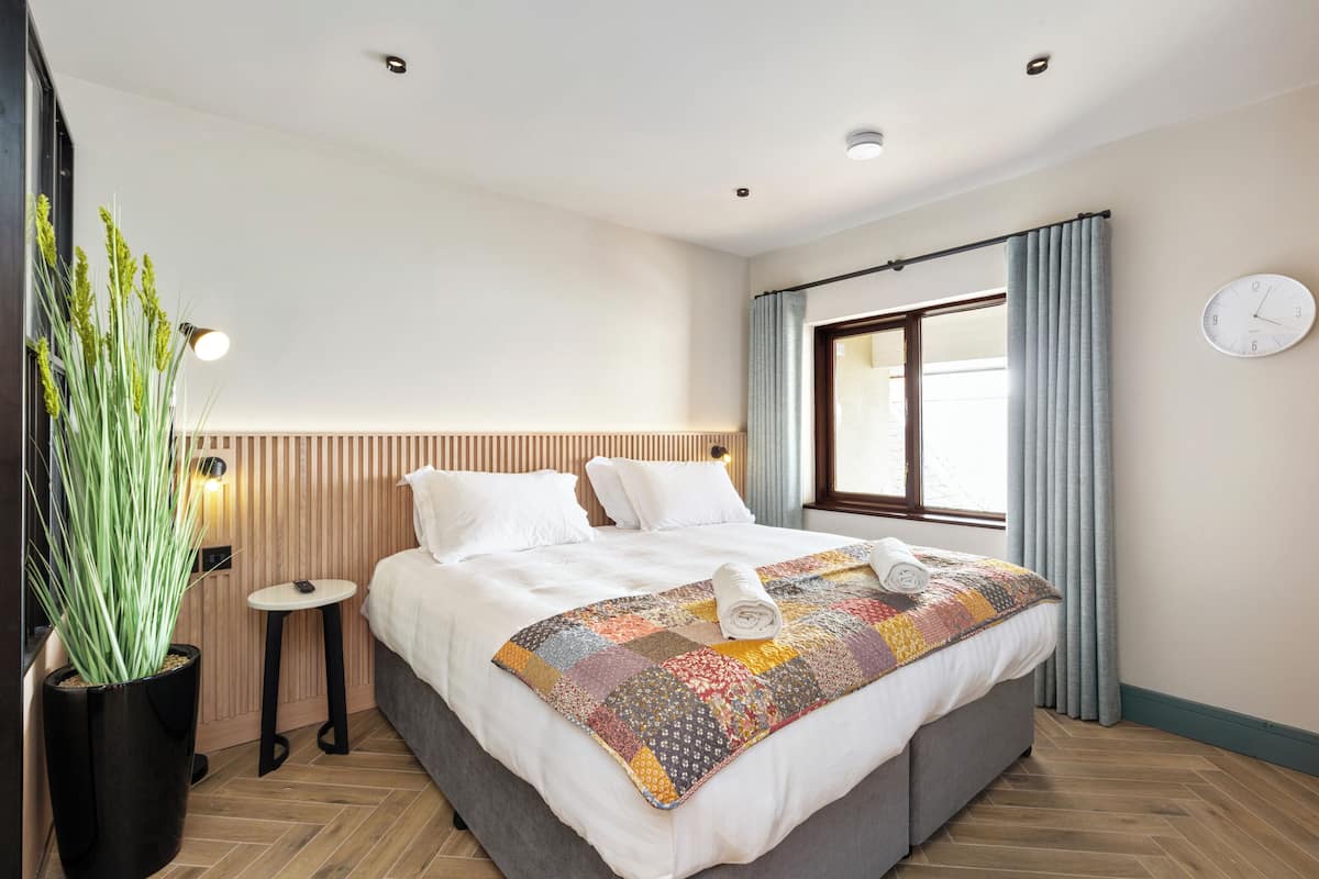 Un lit bien fait, recouvert d’une couverture aux couleurs vives, une plante en pot, une petite table ronde et une horloge murale.