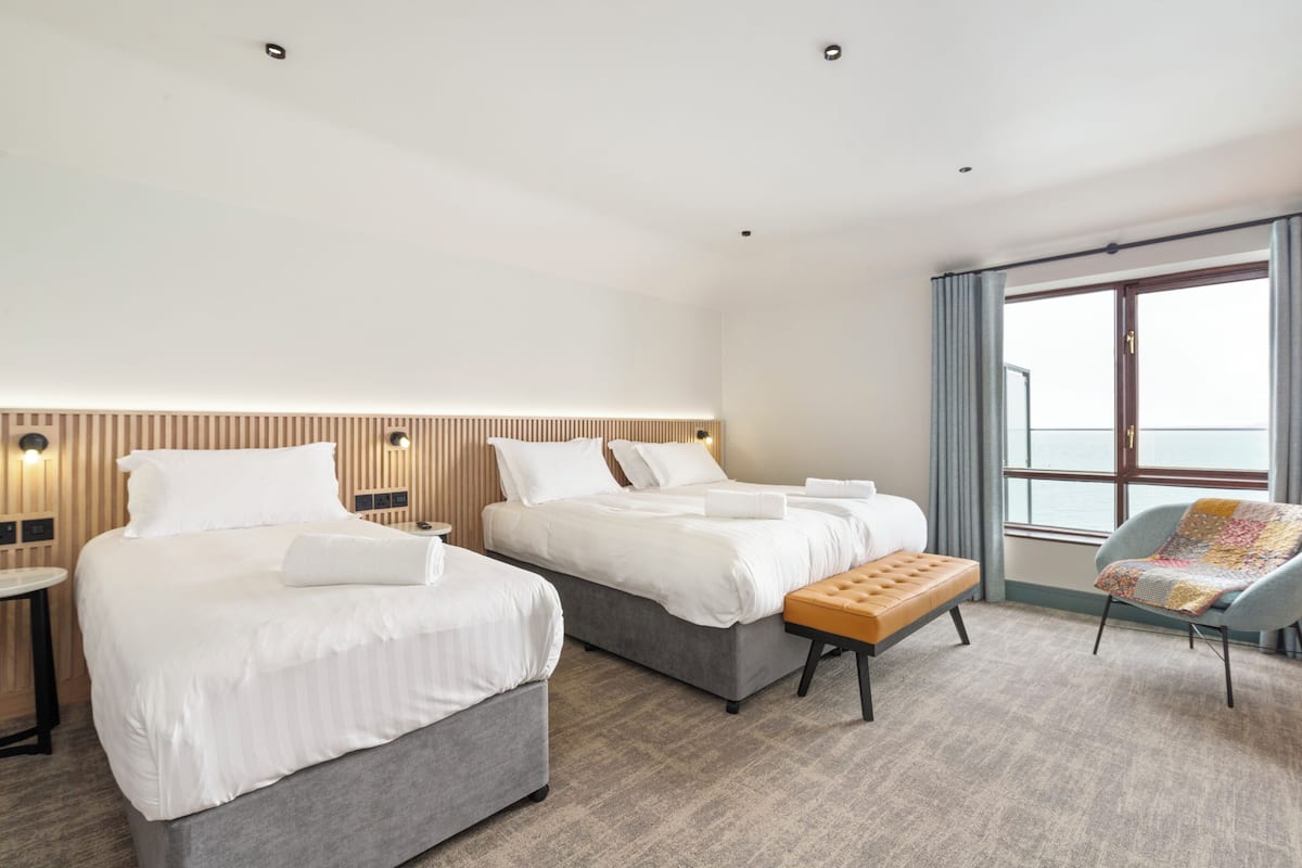 Une chambre d’hôtel avec deux lits, une banquette, une chaise, une petite table et une grande fenêtre avec des rideaux.